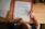 A child is practicing writing the uppercase and lowercase letter 'A' on a worksheet using a black marker. The sheet includes guidelines for tracing the letter. Other worksheets and a box of colored pencils are visible on the wooden table, suggesting an educational environment.