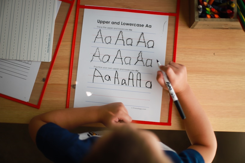 A child is practicing writing the uppercase and lowercase letter 'A' on a worksheet using a black marker. The sheet includes guidelines for tracing the letter. Other worksheets and a box of colored pencils are visible on the wooden table, suggesting an educational environment.