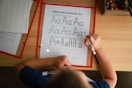 A child is practicing writing the uppercase and lowercase letter 'A' on a worksheet using a black marker. The sheet includes guidelines for tracing the letter. Other worksheets and a box of colored pencils are visible on the wooden table, suggesting an educational environment.