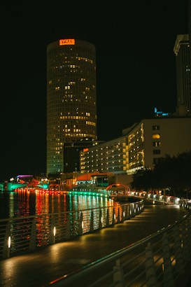 A cityscape at night featuring a tall, cylindrical building with the name SYKES displayed in red lights at the top. The waterfront area below is illuminated by a series of colorful lights reflecting off the water, and a modern railing lines the walkway.