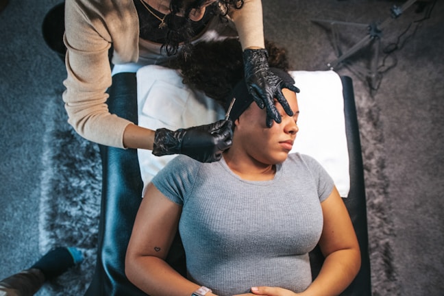 A person lies on a padded table with a gray top, having a beauty or acupuncture treatment. Another person in gloves is delicately performing the treatment, concentrating on the forehead area. The setting is calm, with a textured rug beneath the table and a soft pillow for comfort.
