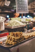 A street food stall featuring trays filled with various fried foods, including spring rolls and crispy-fried items. Behind the trays, there are chopped vegetables like cabbage inside a green basket. A metal pot and a blurred person are visible in the background, indicating a busy cooking environment. The scene is lit with warm tones and soft indoor lighting.