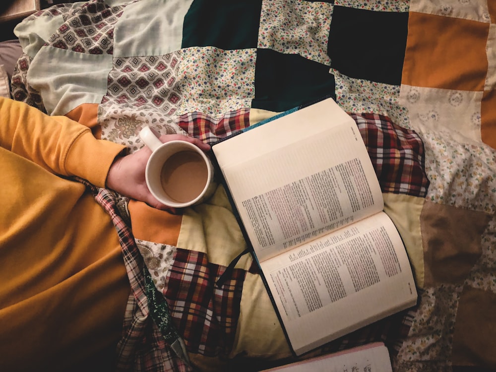 a person laying on a bed with a book and a cup of coffee