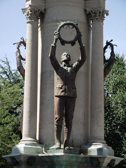 A bronze statue of a man in uniform holding a circular wreath above his head stands prominently against a stone column. The statue is part of a larger monument and other similar statues can be partially seen in the background. The surrounding area includes trees and clear blue skies, conveying a sense of open space and grandeur.