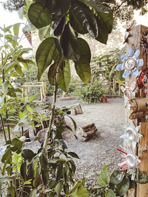 A garden area features a variety of green plants and trees with large leaves. A tree trunk is decorated with flowers made from recycled materials, such as aluminum cans with petals painted in various colors. The ground is covered in gravel and logs are scattered around, contributing to a rustic atmosphere.