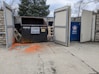 Two large waste containers are inside an open fenced enclosure. One container is black with a cardboard only sign and the other is blue with Republic Services branding. The ground in front is stained with orange paint, and there is a wooden palette and some debris nearby. Trees and stone walls are visible in the background.