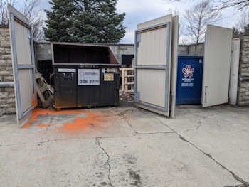 Two large waste containers are inside an open fenced enclosure. One container is black with a cardboard only sign and the other is blue with Republic Services branding. The ground in front is stained with orange paint, and there is a wooden palette and some debris nearby. Trees and stone walls are visible in the background.