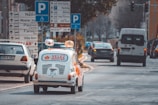A retro-style white car with red branding, driving down a city street lined with multiple road signs and other vehicles. The street is bustling with traffic, and the surrounding area includes buildings and greenery.