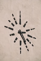 A minimalist clock is mounted on a plain textured wall. The clock face is marked by black rectangular blocks representing the hours, radiating outward in a sunburst pattern. The absence of numbers gives it a modern and abstract aesthetic.