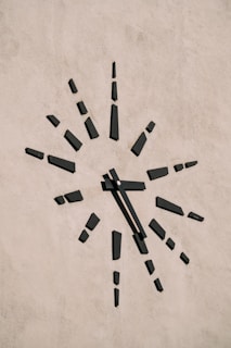 A minimalist clock is mounted on a plain textured wall. The clock face is marked by black rectangular blocks representing the hours, radiating outward in a sunburst pattern. The absence of numbers gives it a modern and abstract aesthetic.