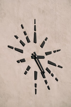 A minimalist clock is mounted on a plain textured wall. The clock face is marked by black rectangular blocks representing the hours, radiating outward in a sunburst pattern. The absence of numbers gives it a modern and abstract aesthetic.
