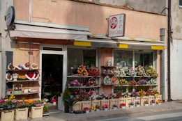 A flower shop with an array of colorful flower arrangements displayed outside. There are baskets and pots of flowers arranged on shelves, each with vibrant blooms. The shop has a large window, and a sign above it features a floral logo.