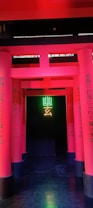 A series of vibrant pink columns with Japanese text create a pathway leading to a dark background. In the center, neon green and yellow symbols illuminate the entrance, adding a modern and mysterious vibe.