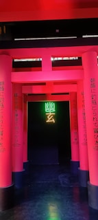 A series of vibrant pink columns with Japanese text create a pathway leading to a dark background. In the center, neon green and yellow symbols illuminate the entrance, adding a modern and mysterious vibe.