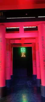 A series of vibrant pink columns with Japanese text create a pathway leading to a dark background. In the center, neon green and yellow symbols illuminate the entrance, adding a modern and mysterious vibe.