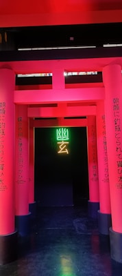 A series of vibrant pink columns with Japanese text create a pathway leading to a dark background. In the center, neon green and yellow symbols illuminate the entrance, adding a modern and mysterious vibe.