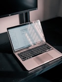 A silver laptop displaying a digital document titled '21 Day Challenge' sits on a dark desk. The screen reflects a grid-like pattern of sunlight and shadows. Behind it, part of a black computer monitor is visible, and the walls appear neutral in color.