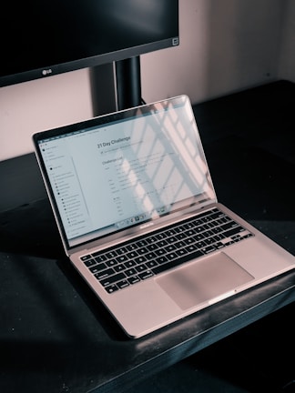 A silver laptop displaying a digital document titled '21 Day Challenge' sits on a dark desk. The screen reflects a grid-like pattern of sunlight and shadows. Behind it, part of a black computer monitor is visible, and the walls appear neutral in color.