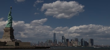 A panoramic view featuring the Statue of Liberty on the left, standing tall against a backdrop of fluffy clouds and a clear blue sky. In the distance, the New York City skyline is visible, including notable skyscrapers and urban structures. The ocean in the foreground reflects the sky.