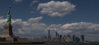 A panoramic view featuring the Statue of Liberty on the left, standing tall against a backdrop of fluffy clouds and a clear blue sky. In the distance, the New York City skyline is visible, including notable skyscrapers and urban structures. The ocean in the foreground reflects the sky.