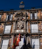 The image features an ornate building facade with detailed classical architecture, including decorative carvings and symmetrical window placements. Bright murals depicting human figures adorn the structure. A sign labeled 'Plaza Mayor' is positioned prominently, accompanied by several colorful flags below. The sky is a clear blue, providing a vibrant backdrop.