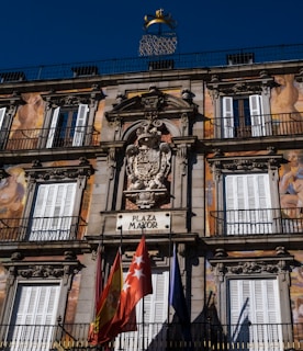The image features an ornate building facade with detailed classical architecture, including decorative carvings and symmetrical window placements. Bright murals depicting human figures adorn the structure. A sign labeled 'Plaza Mayor' is positioned prominently, accompanied by several colorful flags below. The sky is a clear blue, providing a vibrant backdrop.