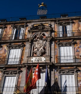 The image features an ornate building facade with detailed classical architecture, including decorative carvings and symmetrical window placements. Bright murals depicting human figures adorn the structure. A sign labeled 'Plaza Mayor' is positioned prominently, accompanied by several colorful flags below. The sky is a clear blue, providing a vibrant backdrop.