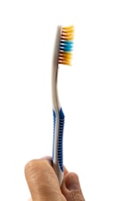 A hand is holding a toothbrush with blue and yellow bristles against a white background. The handle of the toothbrush has blue grips, and the bristles appear clean and unused.