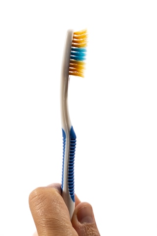 A hand is holding a toothbrush with blue and yellow bristles against a white background. The handle of the toothbrush has blue grips, and the bristles appear clean and unused.
