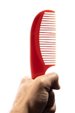 A hand holding a bright red plastic comb with a curved handle, set against a plain white background.
