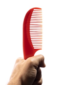 A hand holding a bright red plastic comb with a curved handle, set against a plain white background.