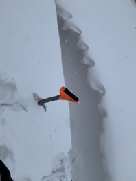 A snow-covered landscape with a deep, vertical snow trench. An ice axe with an orange handle is stuck vertically into the trench wall.