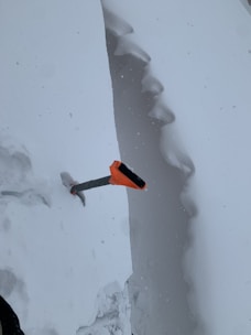 A snow-covered landscape with a deep, vertical snow trench. An ice axe with an orange handle is stuck vertically into the trench wall.
