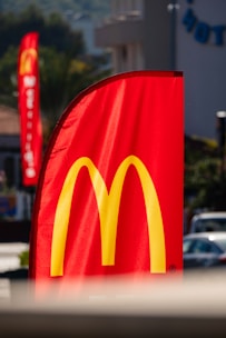 A tall red flag prominently displays the yellow arch logo of a fast-food chain. The flag is situated outdoors with blurred background elements, including another similar flag and buildings. The sunlight highlights the vibrant colors.
