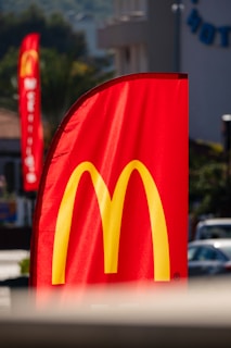 A tall red flag prominently displays the yellow arch logo of a fast-food chain. The flag is situated outdoors with blurred background elements, including another similar flag and buildings. The sunlight highlights the vibrant colors.