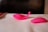 A brightly colored, pink silicone object rests on a white surface. The object has smooth, curved lines and a sleek, modern design. Another similar object is partially visible on the right side. The background is slightly blurred, creating a focus on the objects.