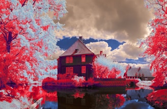A surreal, infrared landscape featuring a house by a reflective body of water surrounded by vibrant red foliage. The sky is filled with dramatic, fluffy clouds that contrast with the blue sky visible in patches.
