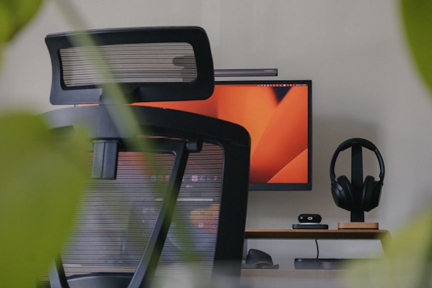 An office setup with a modern ergonomic chair in the foreground, a computer monitor displaying an abstract orange design, a sleek desk with minimal accessories like headphones on a stand, a webcam, and a compact computer or hub. The image is framed by green leaves, suggesting a calm work environment with a touch of nature.