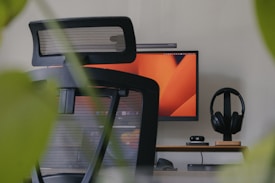 An office setup with a modern ergonomic chair in the foreground, a computer monitor displaying an abstract orange design, a sleek desk with minimal accessories like headphones on a stand, a webcam, and a compact computer or hub. The image is framed by green leaves, suggesting a calm work environment with a touch of nature.