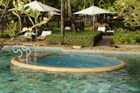 A serene outdoor pool with a circular jacuzzi section surrounded by green foliage and palm trees. There are white lounge chairs and umbrellas on the pool deck, and a pathway lined with manicured bushes.