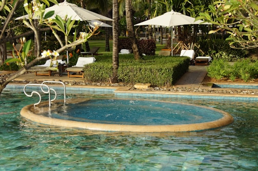 A serene outdoor pool with a circular jacuzzi section surrounded by green foliage and palm trees. There are white lounge chairs and umbrellas on the pool deck, and a pathway lined with manicured bushes.