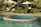 A serene outdoor pool with a circular jacuzzi section surrounded by green foliage and palm trees. There are white lounge chairs and umbrellas on the pool deck, and a pathway lined with manicured bushes.