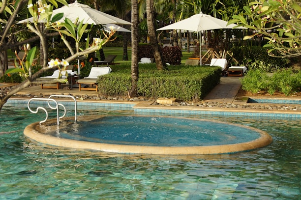 A serene outdoor pool with a circular jacuzzi section surrounded by green foliage and palm trees. There are white lounge chairs and umbrellas on the pool deck, and a pathway lined with manicured bushes.