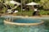 A serene outdoor pool with a circular jacuzzi section surrounded by green foliage and palm trees. There are white lounge chairs and umbrellas on the pool deck, and a pathway lined with manicured bushes.