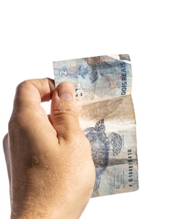 A close-up of a hand holding a boleto payment slip with a laptop in the background.