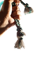 A hand firmly grips a braided rope with tasseled ends. The rope is a mix of turquoise and beige colors, and the light highlights the texture and fibers.