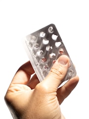 A hand is holding an empty blister pack with multiple circular compartments. The packaging is made of transparent plastic and silver foil, and it appears to be missing several tablets.
