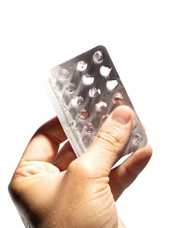 A hand is holding an empty blister pack with multiple circular compartments. The packaging is made of transparent plastic and silver foil, and it appears to be missing several tablets.