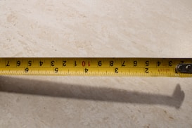 A yellow tape measure is extended across a light-colored surface. The tape displays both metric and imperial measurements. Numbers and markings are clearly visible, with shadows cast on the background.