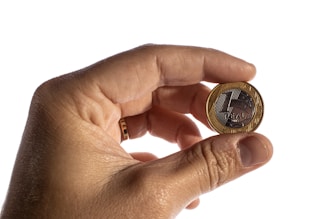 A hand is holding a Brazilian one real coin between the thumb and index finger. The coin features a metallic design with a mix of silver and gold colors, and the words '1 REAL' are visible.